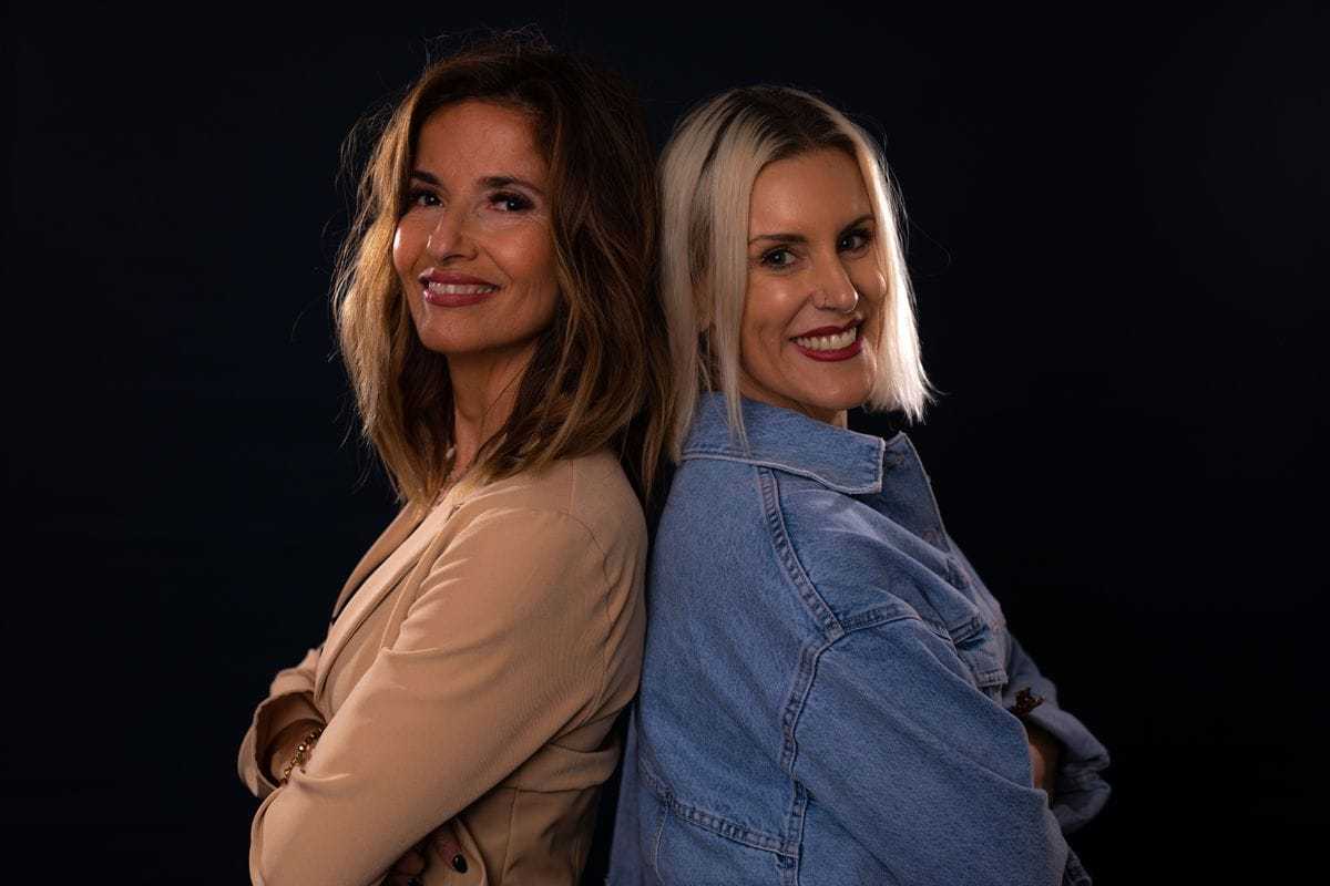 Two women smiling back-to-back against a dark background.