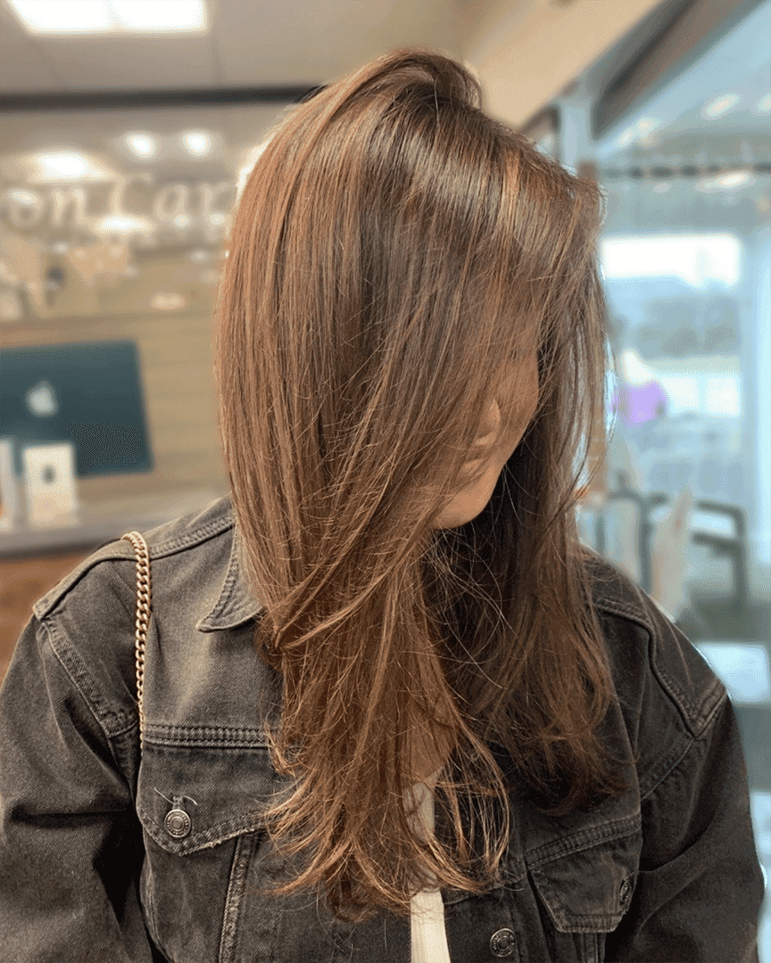 Woman with long, layered brown hair looking down, wearing a denim jacket in a salon.
